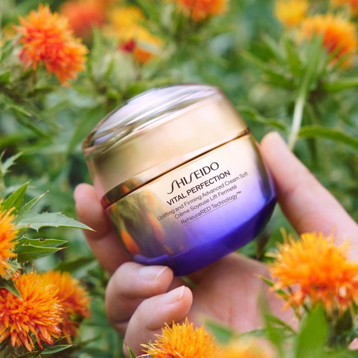 Shiseido Vital Perfection 50 ml uppstramande ansiktslyftkräm i guld- och lila burk som hålls bland blommor