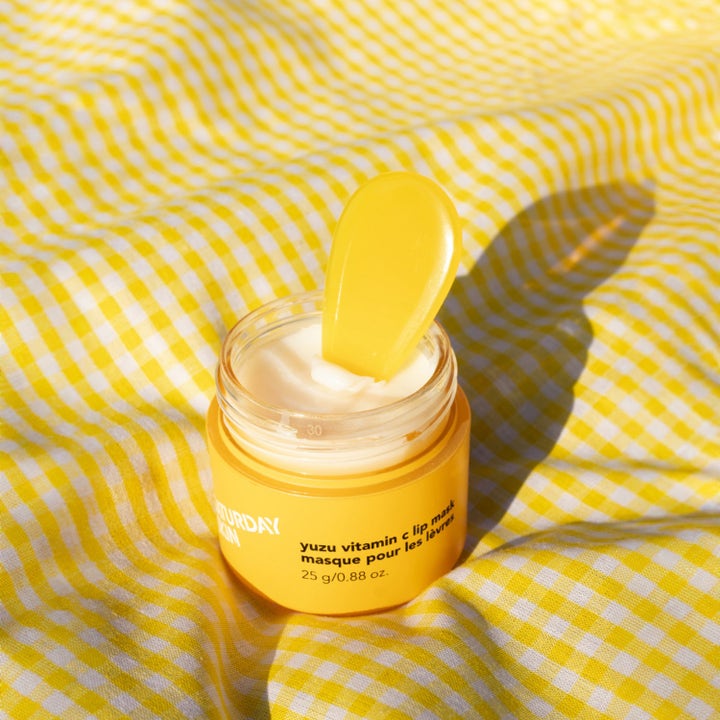 Saturday Skin Yuzu Vitamin C Lip Mask 25 g lystergivande läppmask i gul burk med vit kräm och spatel på gult tyg