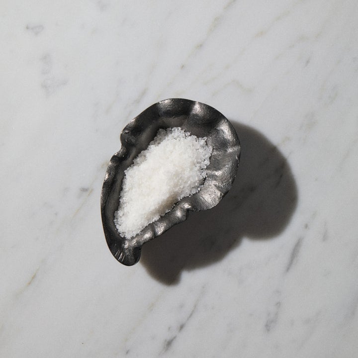 Bodyologist Calming Crystals 250 g lugnande badsalt med vita kristaller i en mörk snäckformad skål på marmor.