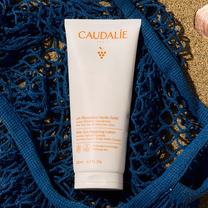 Caudalie Vinosun After-Sun Repairing Lotion 200 ml återfuktande after-sun i vit tub på en blå nätkasse i sanden.