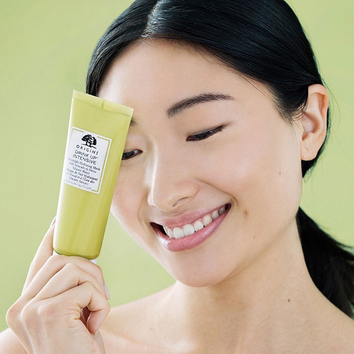 Origins Drink Up Intensive Overnight Mask 75 ml ansiktsmask för djup återfuktning i grön tub som hålls av en kvinna.