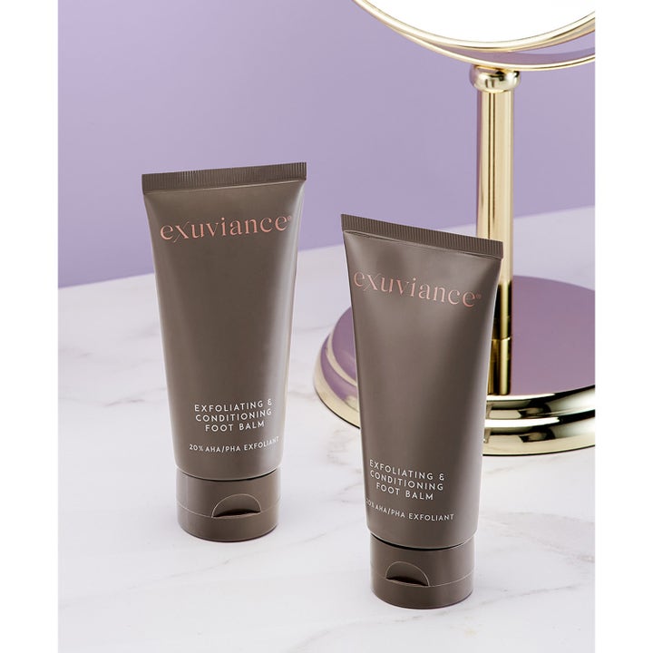 Exuviance Exfoliating & Conditioning Foot Balm 50 g AHA fotexfoliering i två bruna tuber på marmor bredvid en guldspegel.