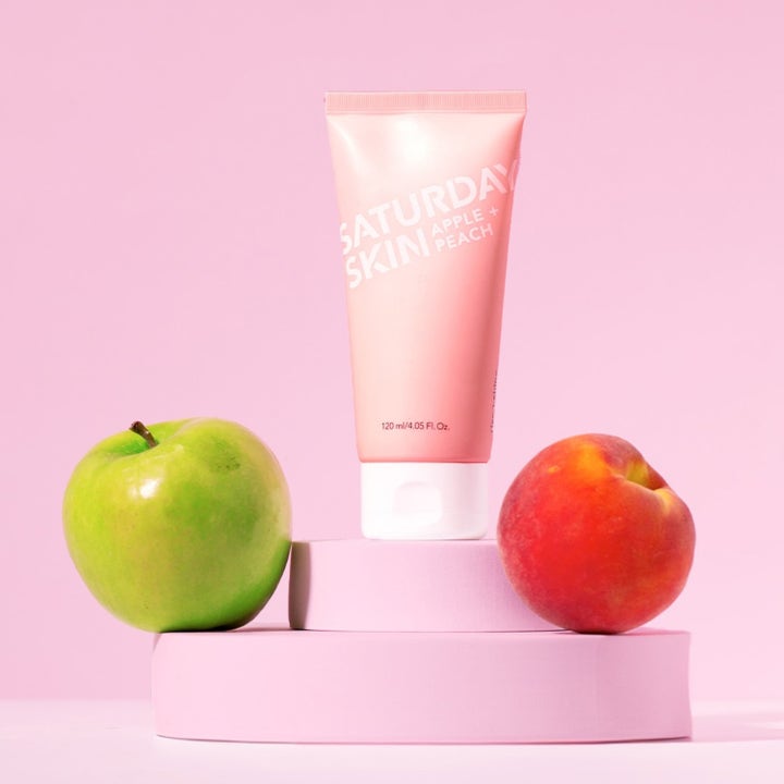 Saturday Skin Rise + Shine Gentle Cleanser 120 ml ansiktsrengöring i rosa tub bredvid ett grönt äpple och en persika.
