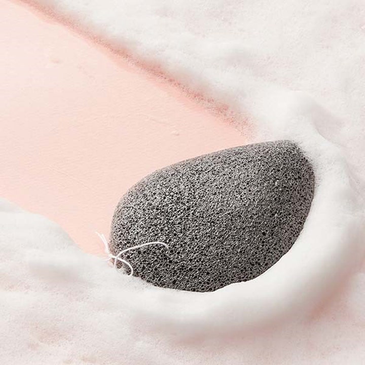 Purito Bamboo Charcoal Konjac Sponge 7 g grå ansiktssvamp med bambuskol omgiven av vitt lödder på rosa bakgrund