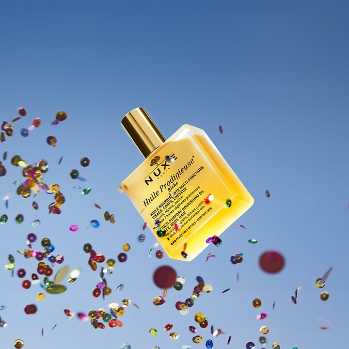 Nuxe Huile Prodigieuse Riche 100 ml fransk återfuktande kroppsolja svävande mot blå himmel med färgglad konfetti.