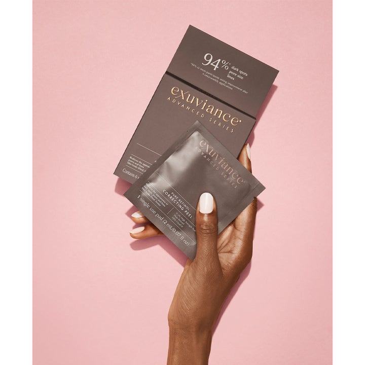Exuviance Pure Retinol Correcting Peel 6 st retinolpeeling i grå sachet som hålls av en hand mot rosa bakgrund.