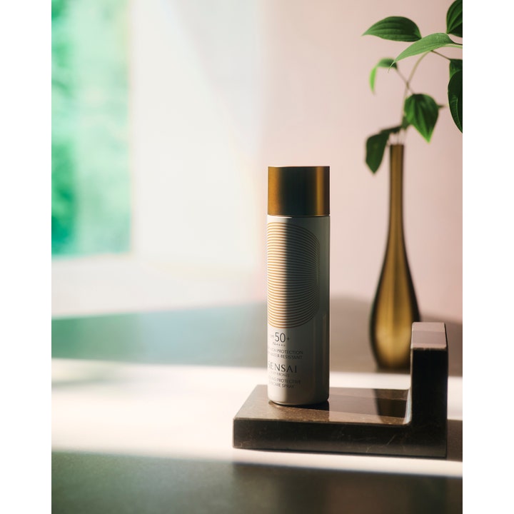 Sensai Silky Bronze Cooling Protective Suncare Spray 150 ml kylande solspray på ett mörkt stenstöd vid en guldvas