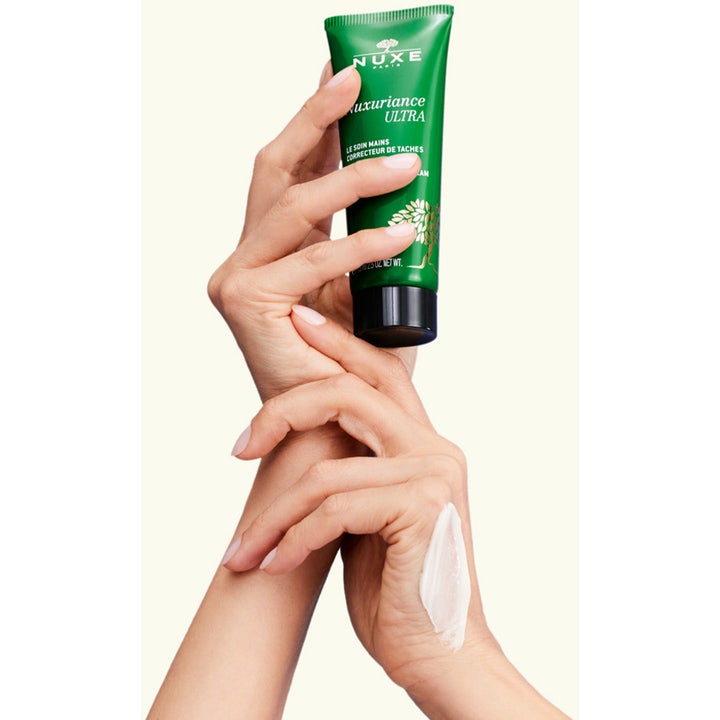 Nuxe Nuxuriance Ultra 90 g anti pigmentering handkräm visas som vit kräm på en hand bredvid den gröna tuben.