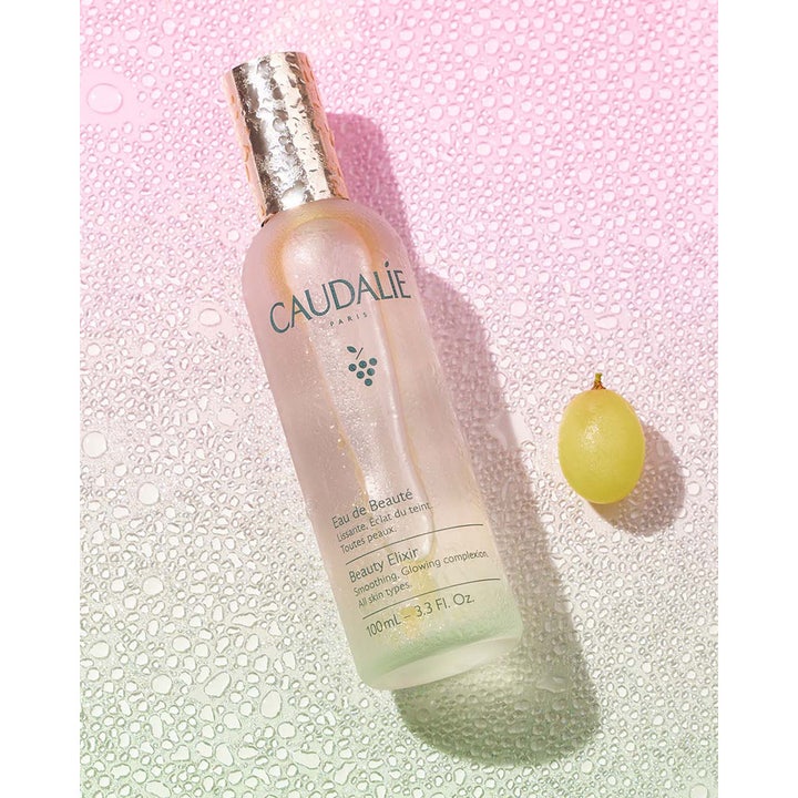 Caudalie Beauty Elixir 100 ml lystergivande ansiktsmist för alla hudtyper i frostat glas bredvid en grön druva
