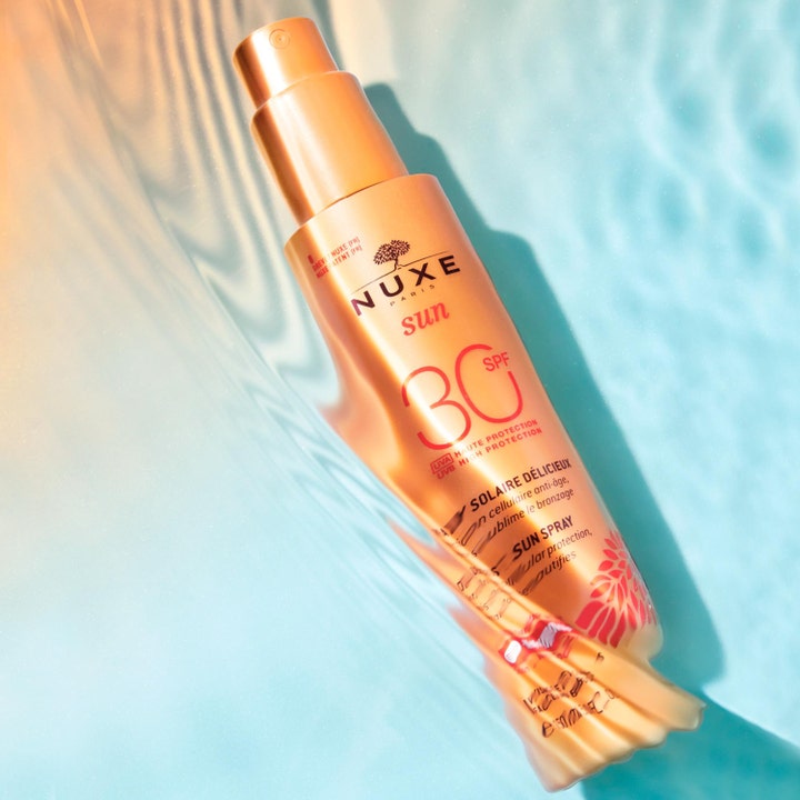 Nuxe Sun Spray SPF30 184 g anti-aging solskydd i en skimrande guldflaska placerad i klart blått vatten