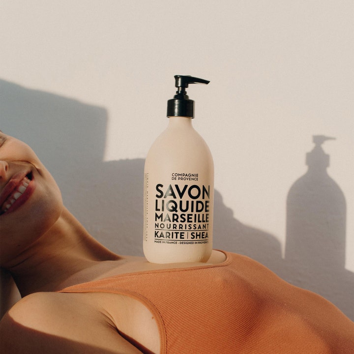 Compagnie de Provence Liquid Marseille Soap 495 ml flytande sheasmör tvål i beige flaska som vilar på en kvinna i solljus
