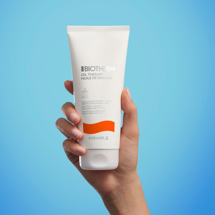 Biotherm Oil Therapy 200 ml mjukgörande duscholja för känslig torr hud i vit tub som hålls i en hand mot blå bakgrund.