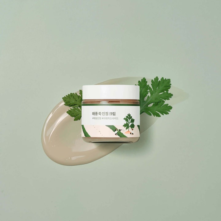 Round Lab Mugwort Calming Cream 80 ml antiinflammatorisk ansiktskräm i burk på mjuk swatch med gröna blad.