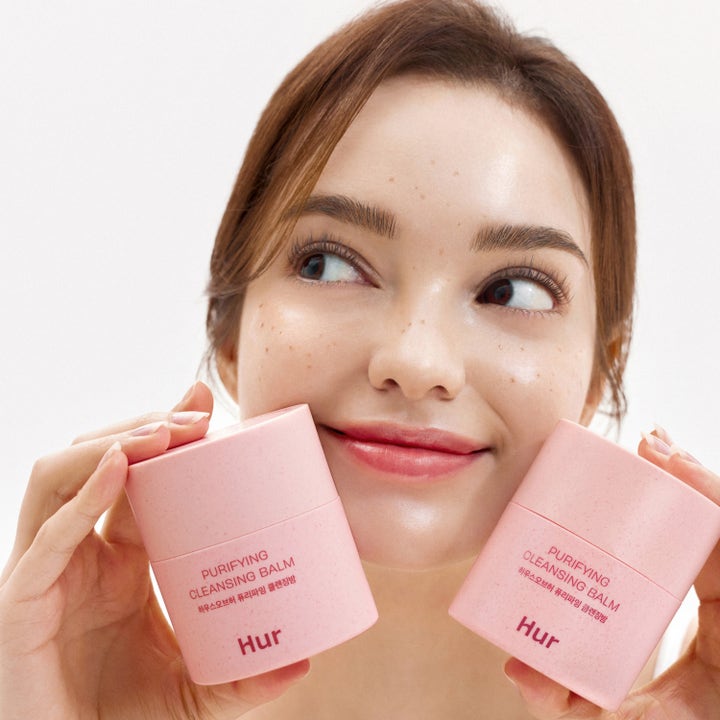House of Hur Purifying Cleansing Balm 50 ml rengöringsbalm i rosa burkar som hålls upp av en leende kvinna.
