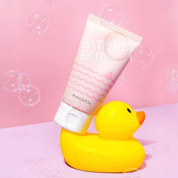 Saturday Skin Rub-a-Dub 75 ml peelinggel på en gul badanka omgiven av såpbubblor för lyster.