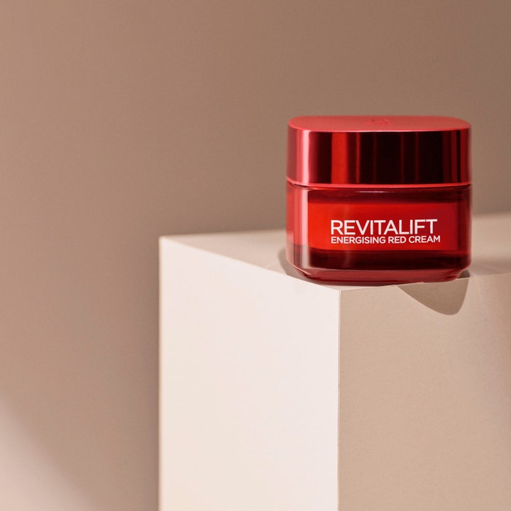 L'Oréal Paris Revitalift Ginseng Glow Day Cream 50 ml anti-age glowkräm i röd glasburk på en ljus beige pelare.