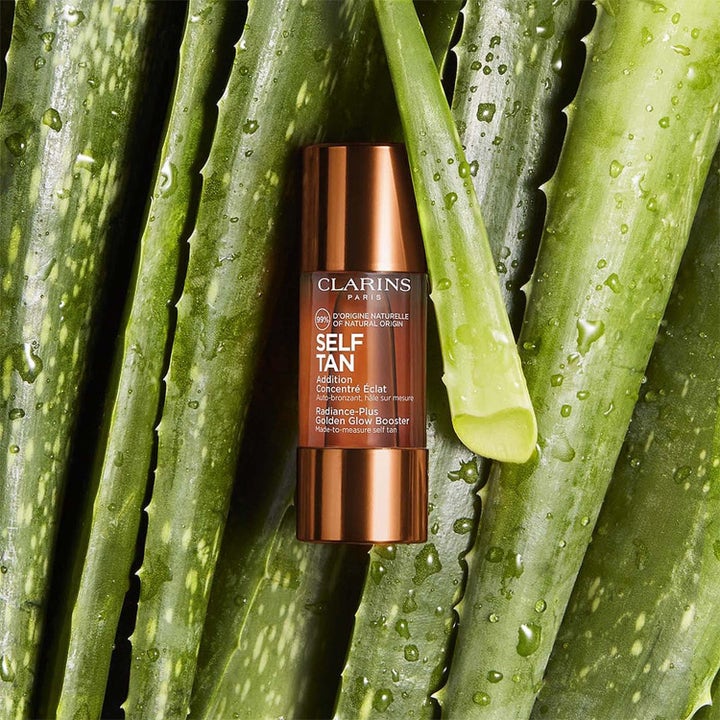 Clarins Golden Glow Booster brun utan sol ger lyster i bronsfärgad flaska bland gröna aloe vera blad med vattendroppar.
