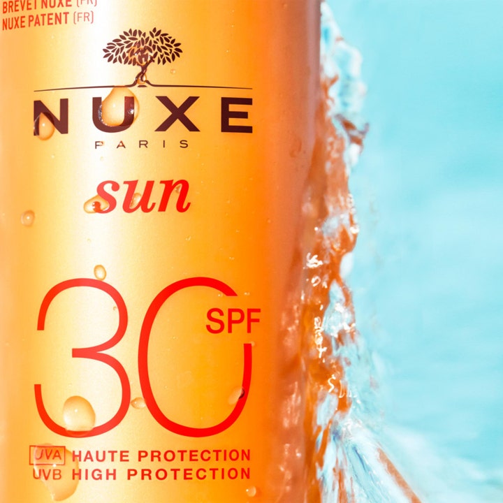 Nuxe Sun Spray SPF30 184 g anti-aging solskydd i orange flaska med friskt stänkande vatten mot en blå bakgrund.