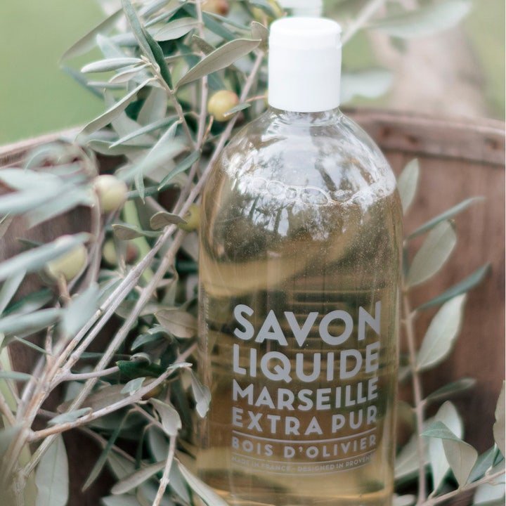 Compagnie de Provence Olive Wood 1000 ml flytande marseille tvål refill i klar flaska bland naturliga olivkvistar.