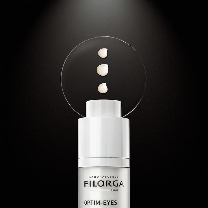Filorga Optim-Eyes 15 ml 3-i-1 ögonkräm mot mörka ringar visar vit flaska och tre droppar krämig textur på glas.