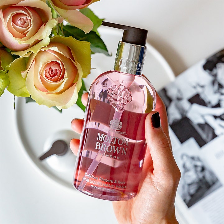 Molton Brown Delicious Rhubarb & Rose 300 ml rengörande handtvål i genomskinlig flaska som hålls bredvid rosor.