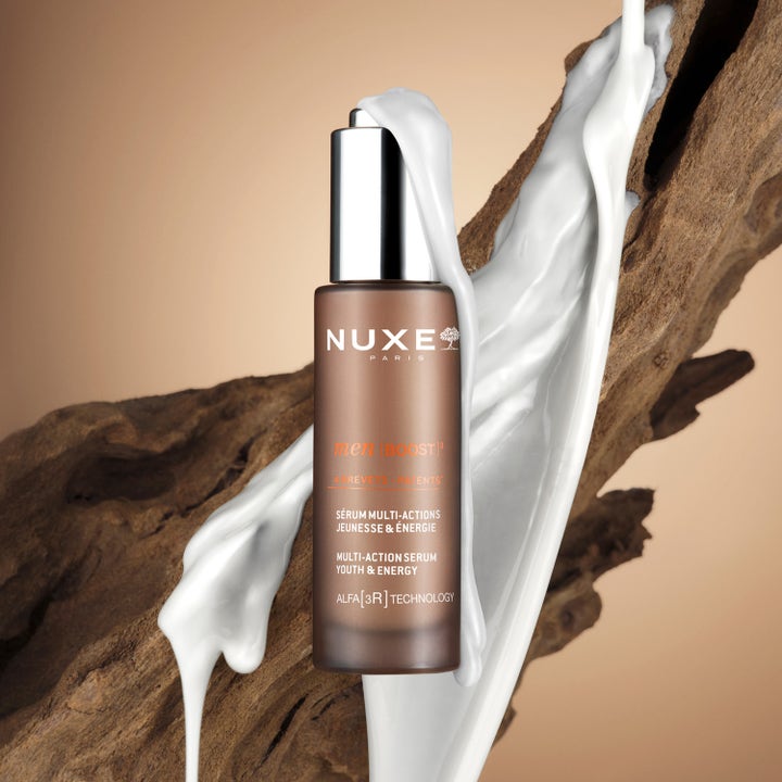 Nuxe Men Multi Action Serum 30 ml ger energi i en brun glasflaska framför trä och rinnande vit kräm.