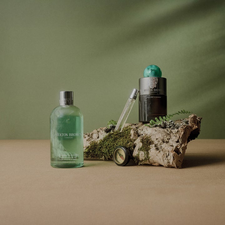 Molton Brown Wild Mint & Lavandin 300 ml uppfriskande duschtvål i grön flaska bredvid parfym och kompass på mossa.