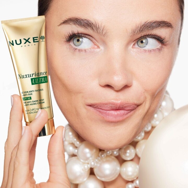 Nuxe Nuxuriance Ultra Anti Dark Spot Cream 50 ml dagkräm i guldtub som hålls av kvinna med stora pärlor