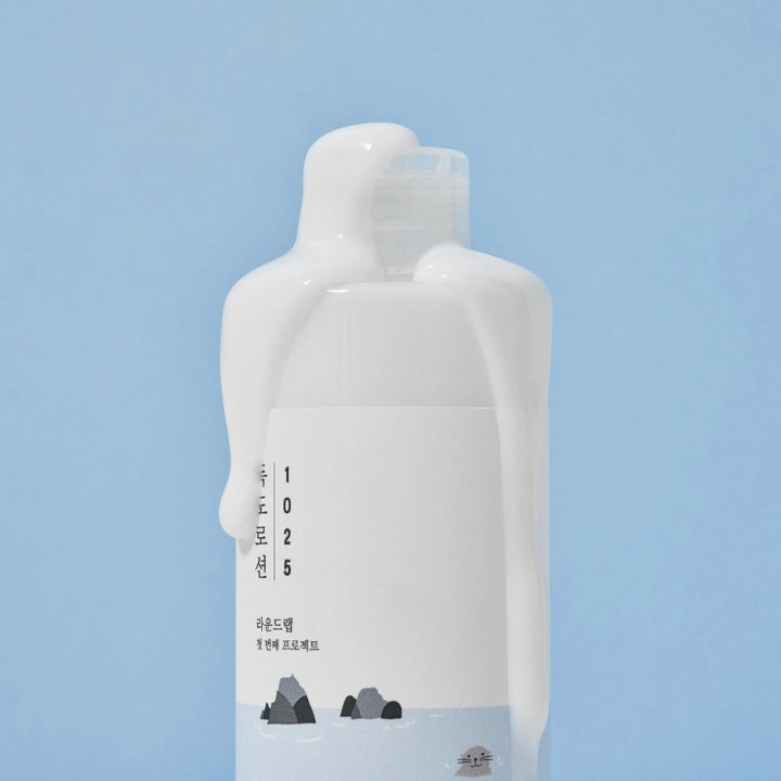 Round Lab 1025 Dokdo Lotion hydrering ansiktslotion med krämig vit formula som rinner nerför flaskan mot blå bakgrund