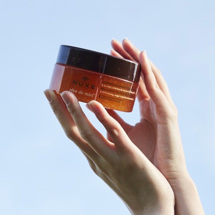 Nuxe Rêve de Miel Body Scrub 175 ml exfoliering för torr hud i en burk som hålls upp mot en klarblå himmel.