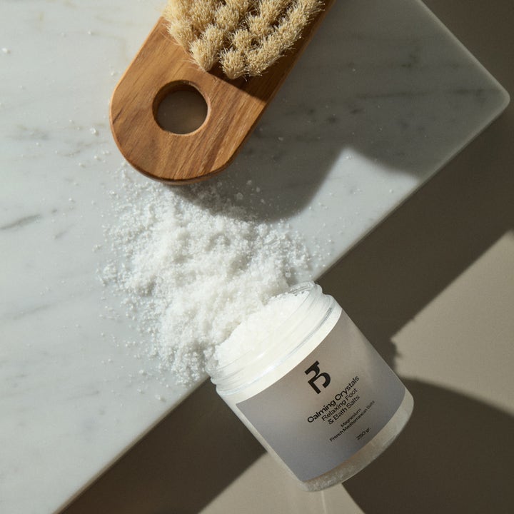 Bodyologist Calming Crystals 250 g lugnande badsalt utspritt på en marmoryta bredvid en träborste.