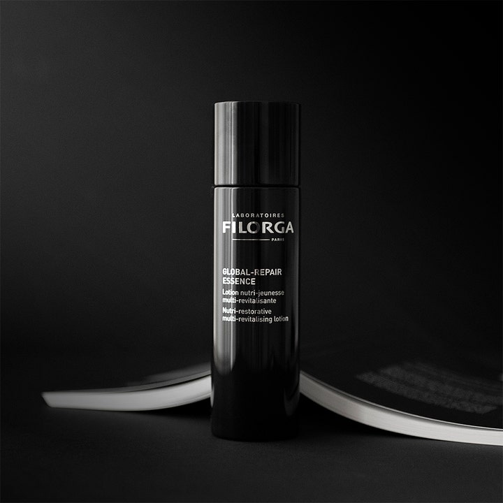 Filorga Global Repair Essence 150 ml anti age essence behandling i en elegant svart flaska intill en öppen bok.