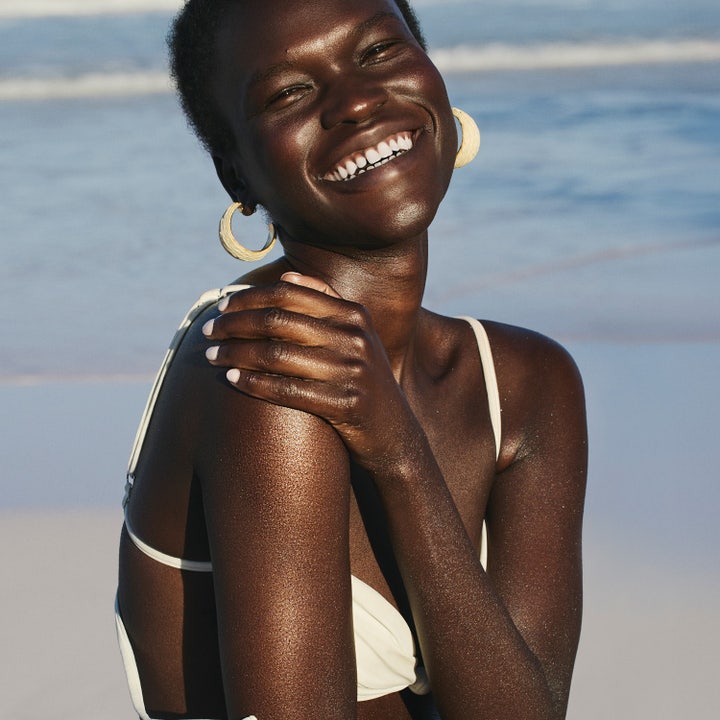 Sol de Janeiro Copacabana Bronze Glow Oil 75 ml ger lyster på en leende kvinna vid en solig strand.