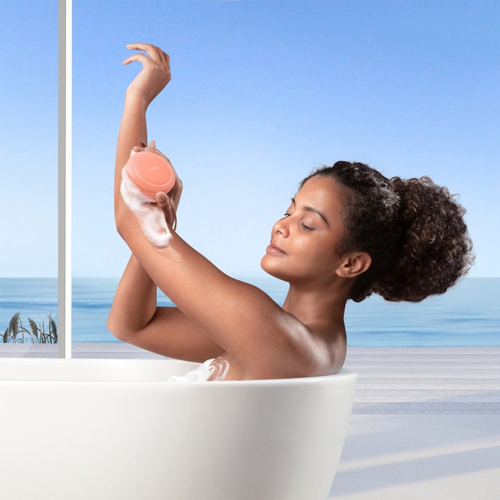 FOREO LUNA 4 body Peach Perfect 1 st kroppsborste ger exfoliering i ett badkar med lödder och vacker havsvy.