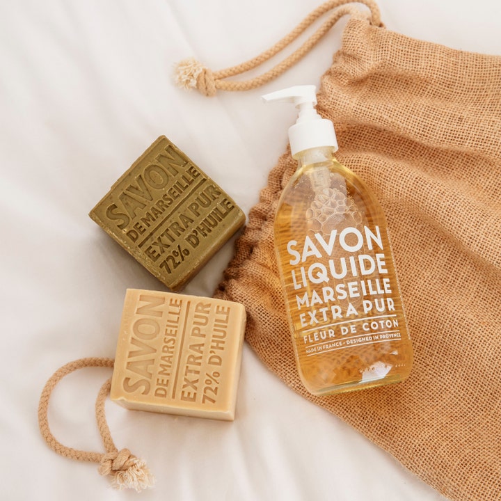 Compagnie de Provence Liquid Marseille Soap 495 ml lyxig handtvål med bomullsblomma bredvid fasta tvålar och en jutepåse.