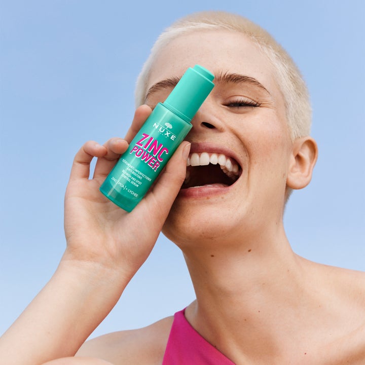 Nuxe Zinc Power Blemish And Spot Control Serum 30 ml mot orenheter i grön flaska som hålls av en person mot blå himmel