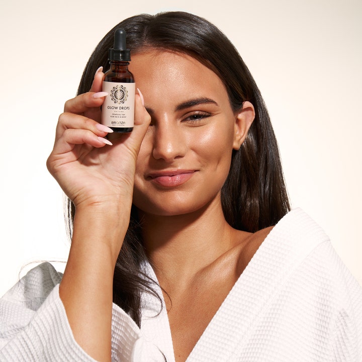 Bronza Glow Drops 30 ml självbrunningsdroppar för naturlig lyster i mörk glasflaska på kvinna med solkysst brun hud.