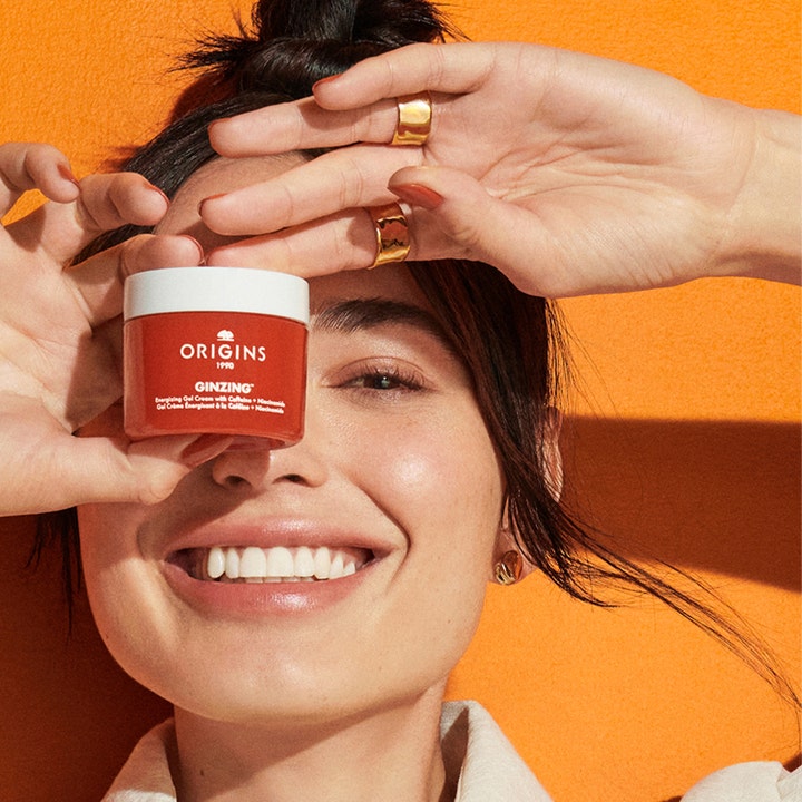 Origins Ginzing Energizing Gel Face Cream 50 ml i orange burk som hålls framför ansiktet på en leende kvinna