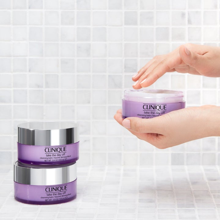 Clinique Take The Day Off 375 ml rengöringsbalm i lila burkar med silverlock som hålls mot vitt kakel.