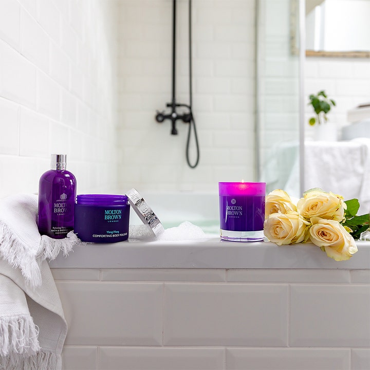 Molton Brown Relaxing Ylang Ylang Bath & Shower Gel 300 ml avkopplande duschtvål i lila flaska med ett tänt ljus.